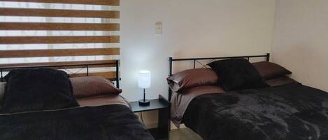 A bedroom with two beds, a nightstand with a lamp, and a window with blinds.