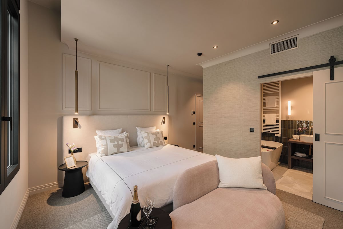 A modern hotel room with a large bed, a bedside table with a bottle and glass, and a bathroom visible through an open door.