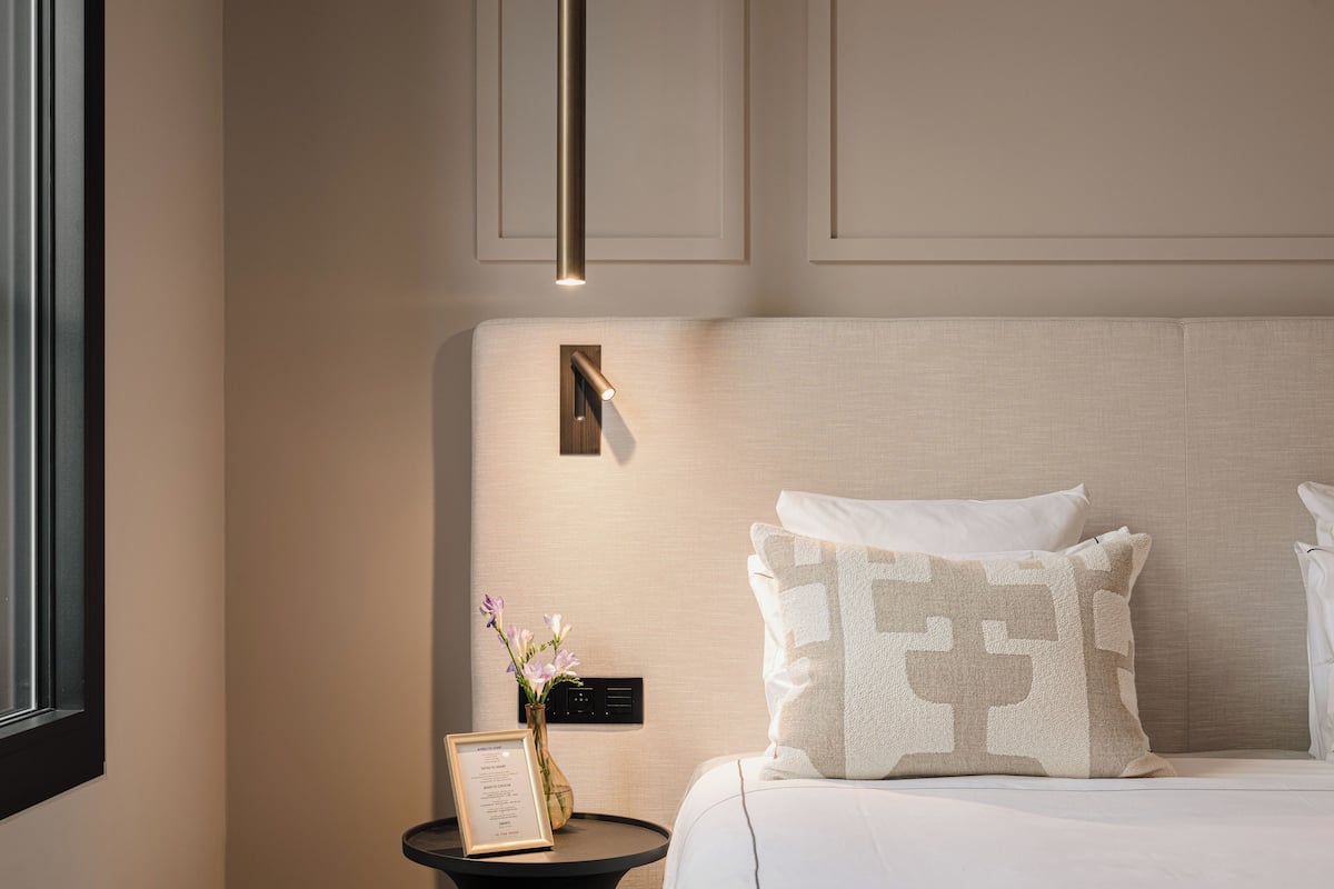 A hotel room with a bed, a bedside table with a vase of flowers, and a wall-mounted control panel.