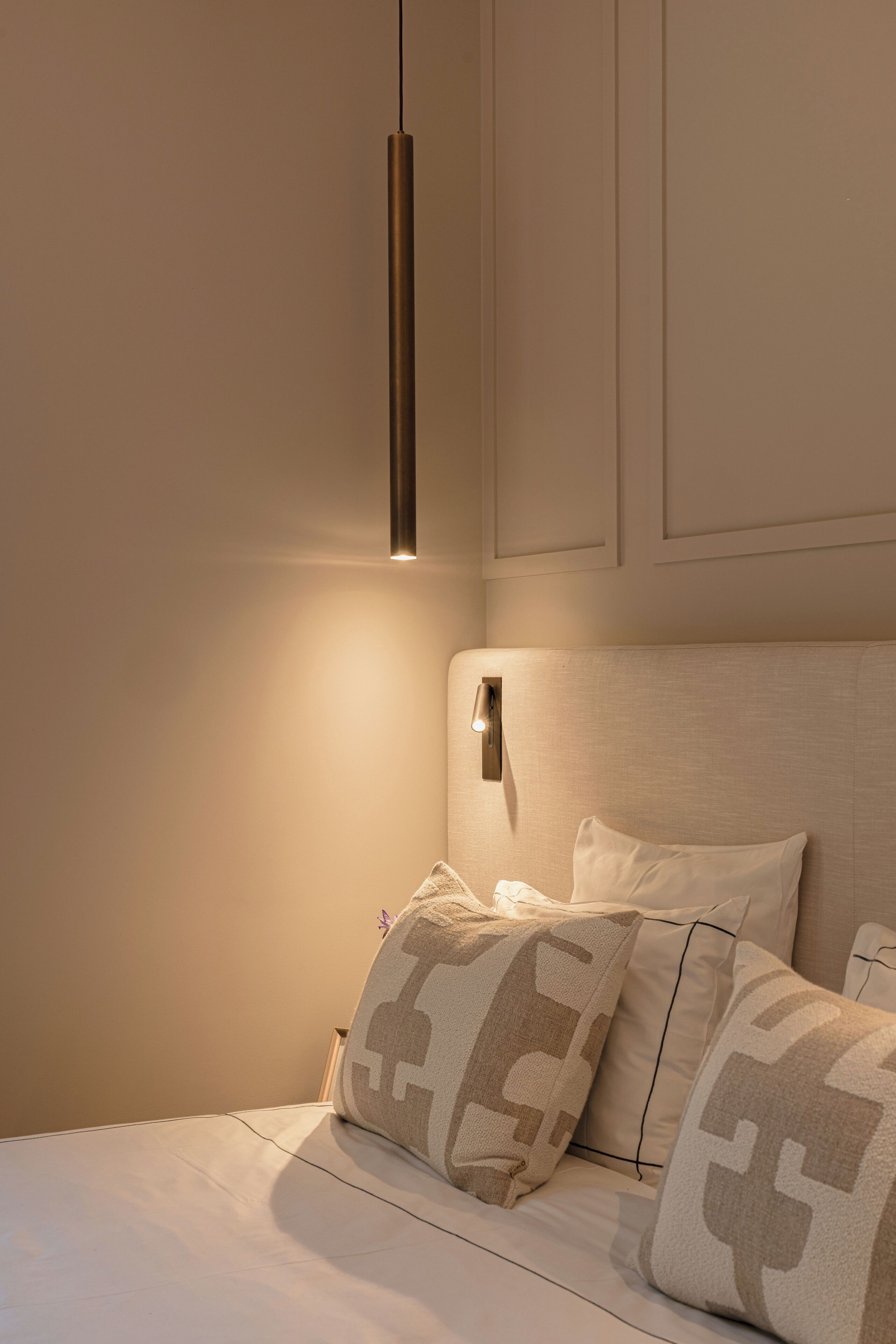 Un lit bien fait, agrémenté d’oreillers à motifs, une lampe de chevet et un luminaire fixé au mur.