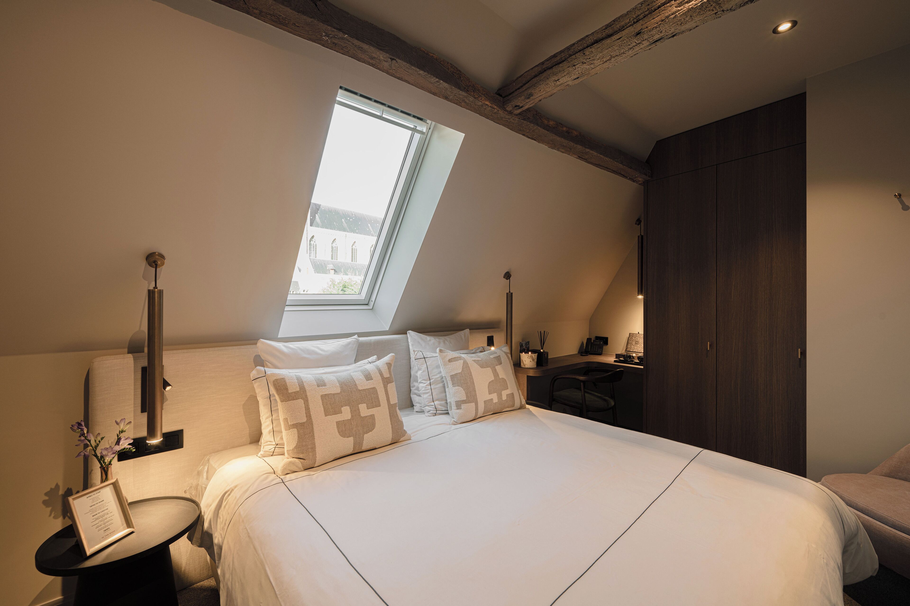 Een slaapkamer met dakraam, een bed met witte beddengoed, een nachtkastje, een lamp en een kledingkast.