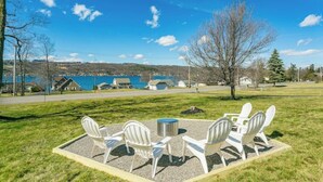 A grassy outdoor seating area with white Adirondack chairs and a metal table, overlooking a body of water and buildings in the background.