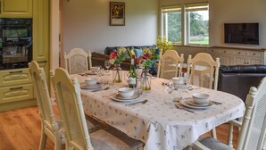 Un comedor con una mesa dispuesta para cuatro personas, rodeada de sillas blancas, una cocina con armarios verdes y un televisor montado en la pared.