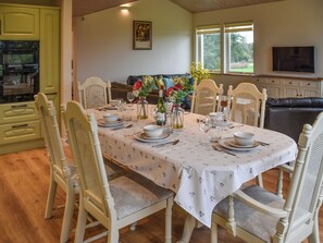 Un comedor con una mesa dispuesta para cuatro personas, rodeada de sillas blancas, una cocina con armarios verdes y un televisor montado en la pared.