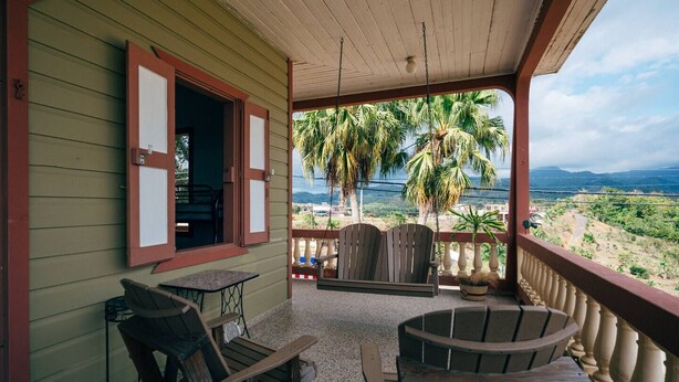 A covered porch with two wooden chairs and a table, overlooking a scenic view with palm trees and mountains. - Antique Mountain Cabin (Ponce)