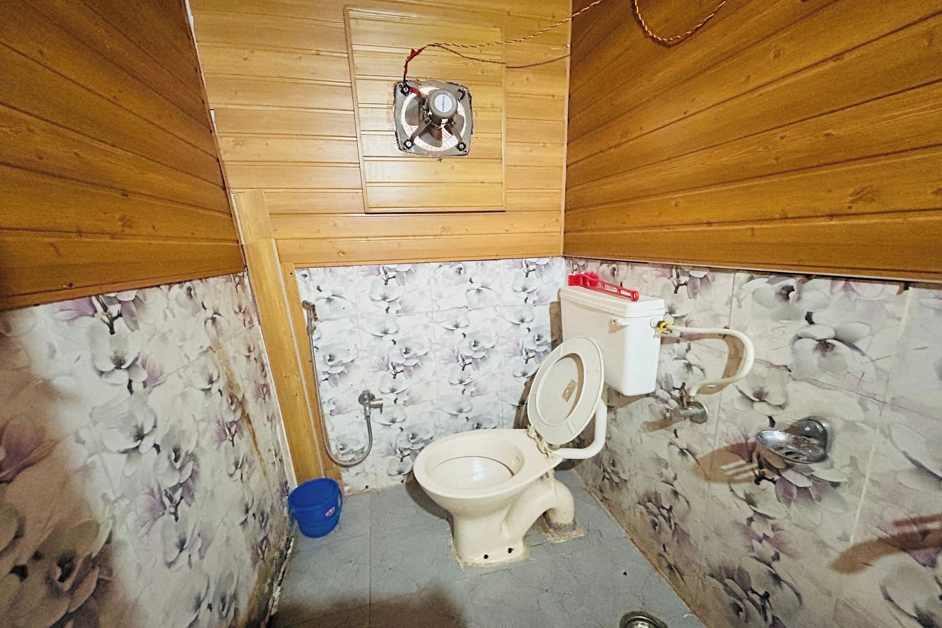 A small, wooden-paneled bathroom with a toilet, a blue bucket, and a shower.