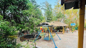 A playground with swings and a slide, surrounded by greenery and a thatched-roof structure. - Rajgad Farms & Agritourism (Pune)