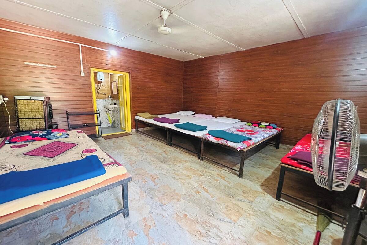 A room with wooden walls, two beds with colorful bedding, a standing heater, a fan, and a tiled floor.