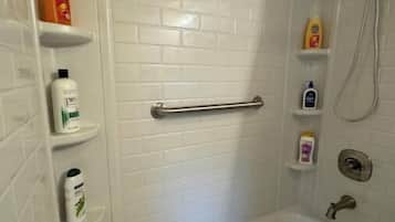 A bathroom with a bathtub, a shower, and a handrail. There are shelves with toiletries.