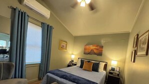 Un dormitorio con cama, mesitas de noche, una silla, un banco, una ventana con cortinas y un cuadro en la pared.
