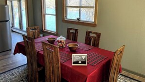 Un área de comedor con mantel rojo, sillas de madera, un cartel que indica la contraseña de WiFi y vistas a un lago a través de las ventanas.