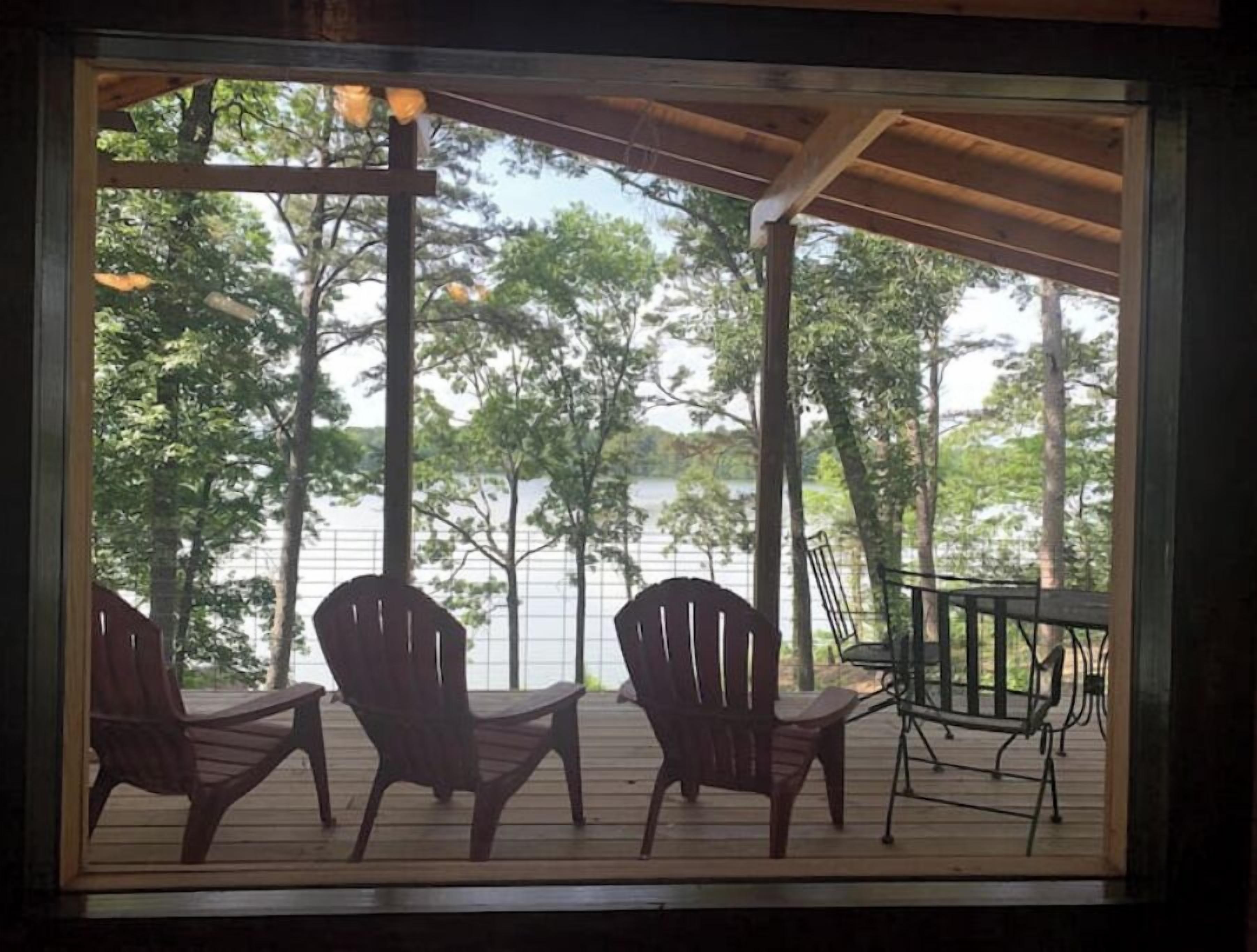 Een houten terras met drie Adirondack-stoelen en een tafel, met uitzicht op water en bomen.