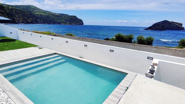 A rectangular pool with a clear blue water surface, surrounded by a white wall and a stone border. In the background, there is a view of the ocean and a rocky island. - Azure Sunset (Mosteiros)