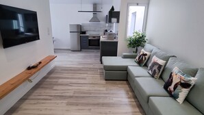 A modern living room with a flat-screen TV, a wooden shelf, a green sofa with decorative pillows, and a kitchen in the background. - Azure Sunset (Mosteiros)