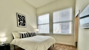 Een slaapkamer met een bed, twee ramen met luiken, een nachtkastje en een aan de muur hangend kunstwerk.