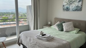 Una habitación de hotel con un ventanal grande, una cama con cubrecama verde, una mesita de noche con lámpara y un cuadro de plantas tropicales en la pared.