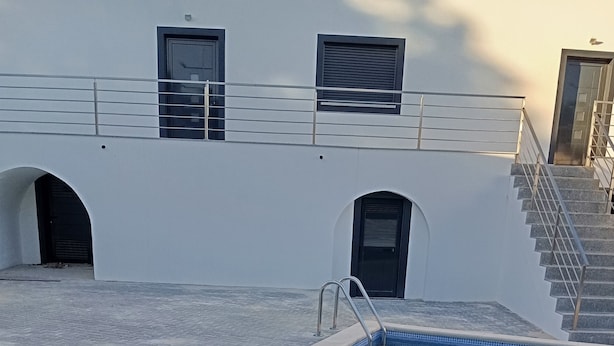 Una piscina con una escalera metálica, borde de azulejos y un edificio con fachada blanca y puertas negras al fondo.
