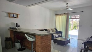 Ein kompakter Wohnraum mit Küchenzeile, die eine Ziegel-Arbeitsplatte und zwei Barhocker hat. Ein Sofa und ein kleiner Tisch befinden sich im Wohnbereich, mit einer Glastür, die zu einem Außenbereich führt.