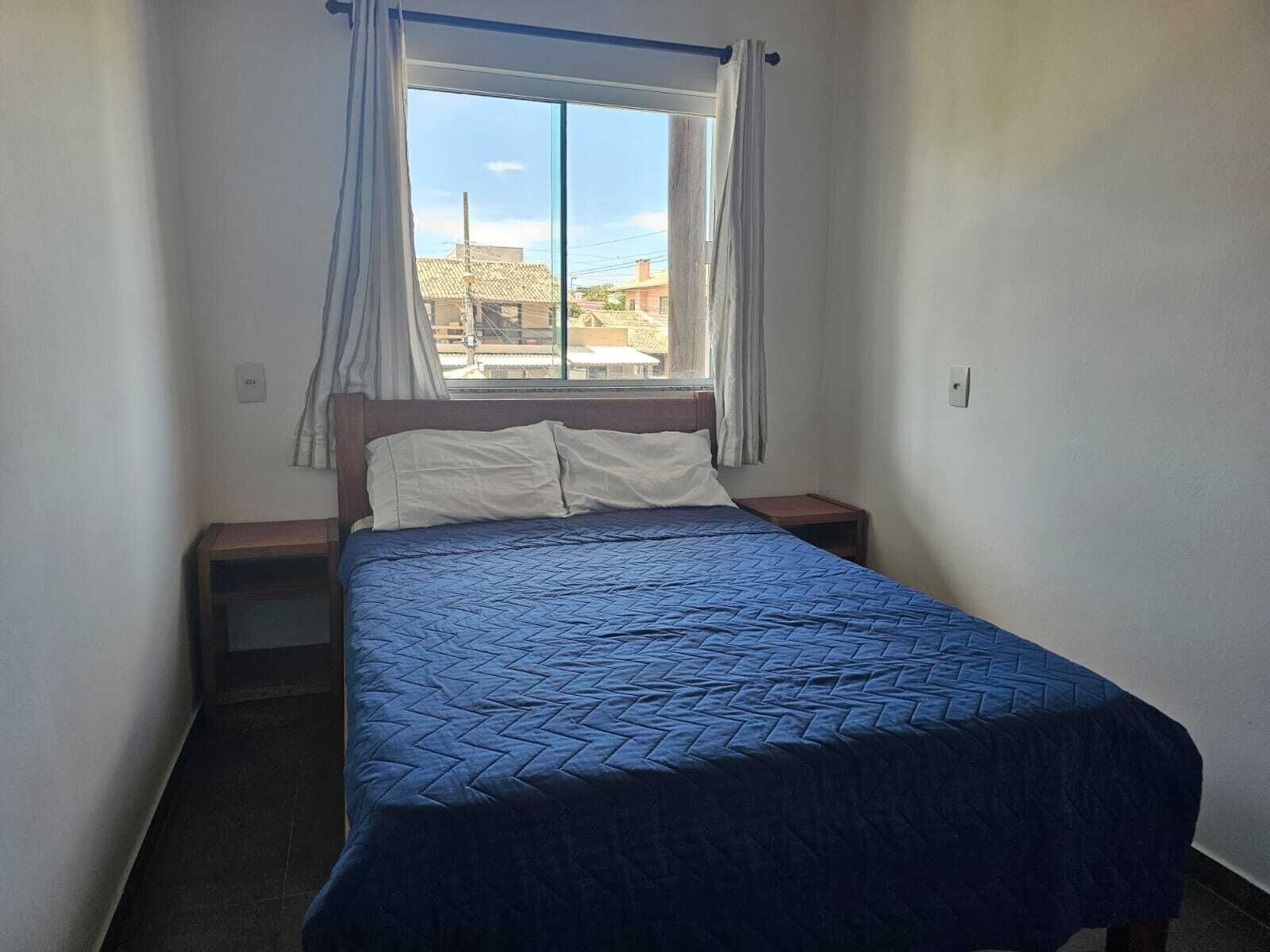 A simple bedroom with a bed, a nightstand, a window with curtains, and a view of buildings outside.