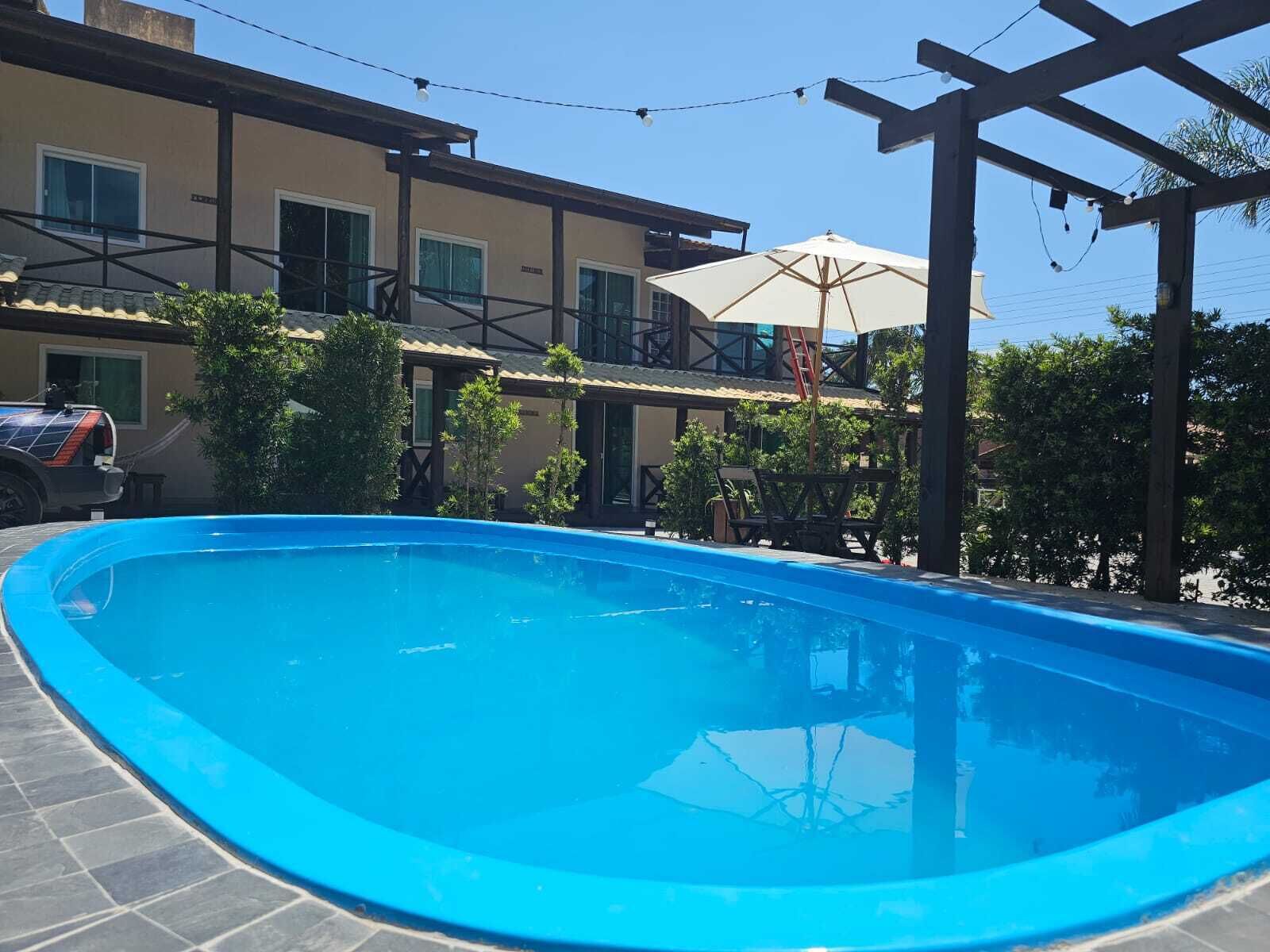 A swimming pool with a pergola and umbrella, surrounded by a building with multiple windows and a car parked nearby.