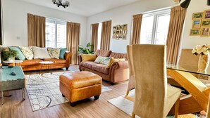 A spacious living room with a leather sofa, a wooden coffee table, and a glass desk. There are large windows with curtains, a chandelier, and framed pictures on the wall. - The Mirrored House (Whole Flat) (Darford)