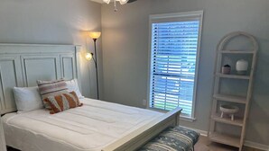 Une chambre à coucher comprenant un lit, un banc, une table de chevet, une lampe, une fenêtre avec des stores et une étagère ornée d’objets décoratifs.