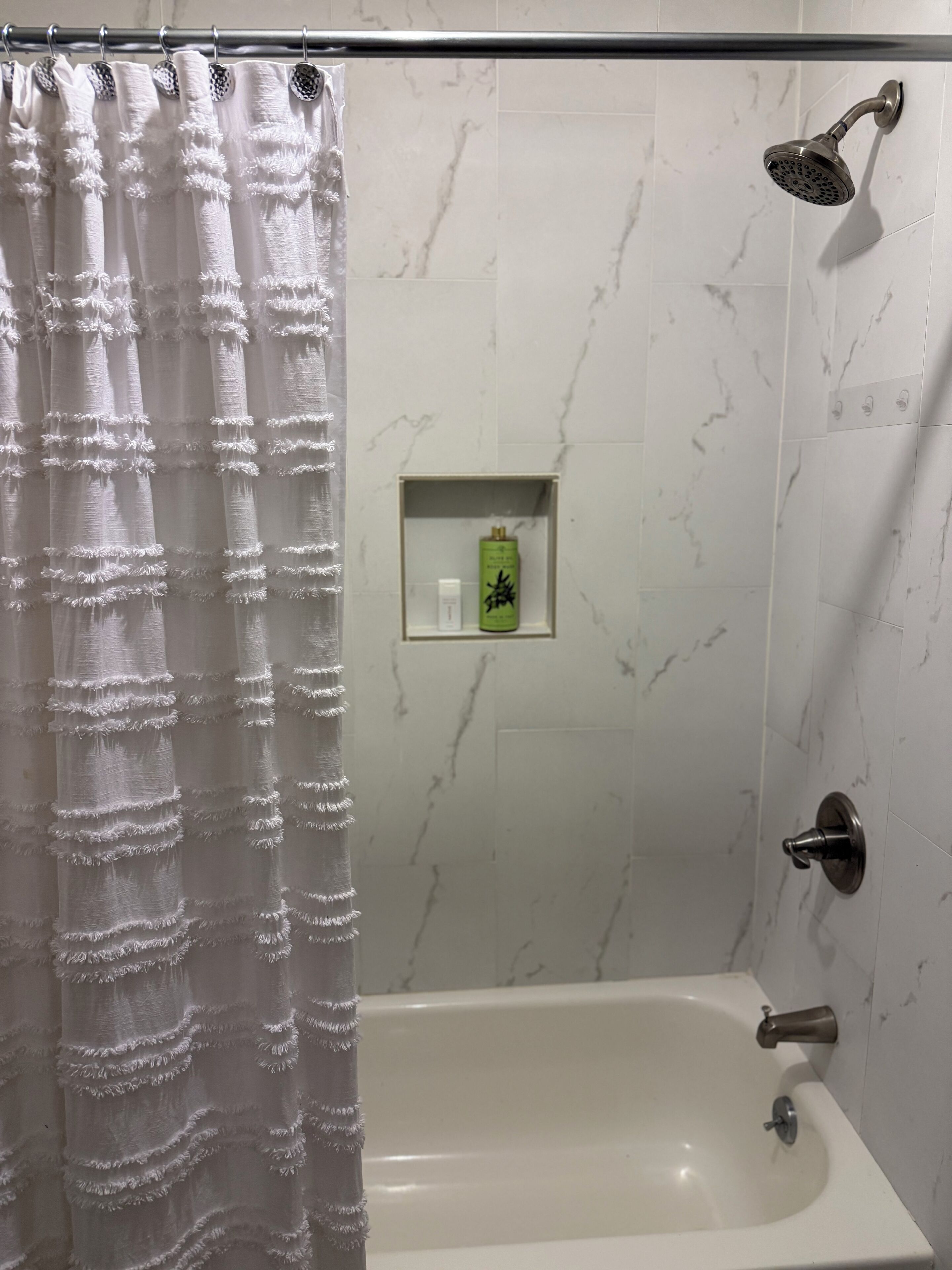 Un baño con ducha, bañera y retrete. Hay una cortina de ducha, una botella verde en la pared y un estante pequeño con artículos de aseo.