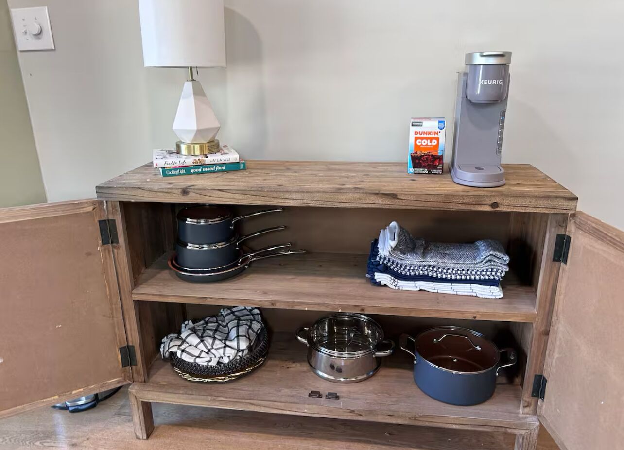 A wooden cabinet with shelves containing kitchenware, a coffee maker, and a box of Dunkin' Cold coffee.
