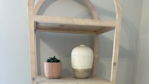 Une étagère en bois à trois niveaux, avec un bol sur l’étagère inférieure, un pot rose contenant une plante sur l’étagère du milieu et un vase blanc sur l’étagère supérieure.