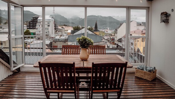 Ein Essbereich mit einem Holztisch und Stühlen, einer Vase mit Blumen und Blick auf Gebäude und Berge durch große Fenster.