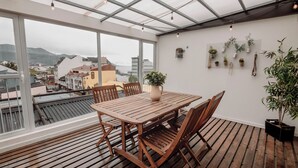 Eine Holzterrasse mit Tisch und Stühlen, einem Blumentopf und Blick auf eine Stadt durch große Fenster.