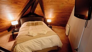 Ein Schlafzimmer mit geneigter Decke, Holzwänden, einem Bett mit weißer Bettwäsche, zwei Nachttischlampen und einem an der Wand montierten Fernseher.