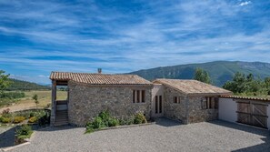 Un bâtiment en pierre avec un toit en tuiles, une allée en gravier et une chaîne de montagnes en arrière-plan.