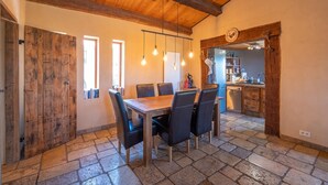 A dining area with a wooden table and chairs, a kitchen in the background, and a stone floor. - Holiday Home "Time-out" with Mountain Views, Shared Pool, and Wi-Fi (Laragne-Montéglin)