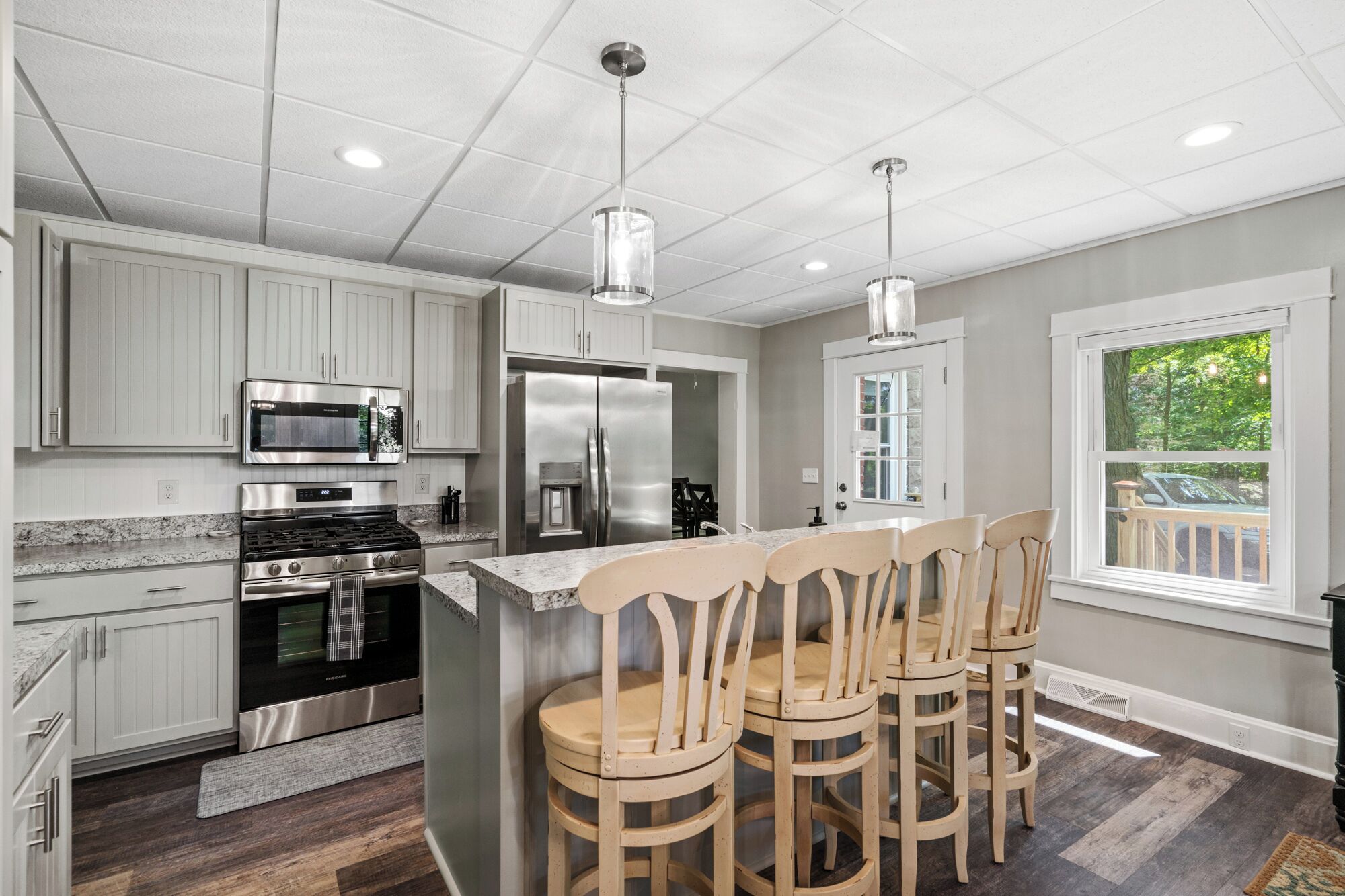 A modern kitchen with white cabinets, stainless steel appliances, a central island with bar stools, and a window with a view of greenery.