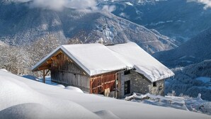 A wooden cabin with a snow-covered roof, set against a backdrop of snow-covered mountains and a clear sky.