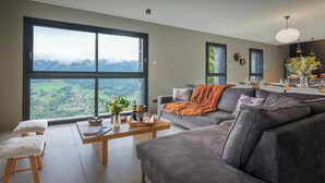 A modern living room with a grey sofa, a wooden coffee table, and a view of a green landscape through large windows.