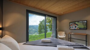 A bedroom with a large bed, a view of a green landscape through a sliding door, a chair, and a flat-screen TV mounted on the wall.