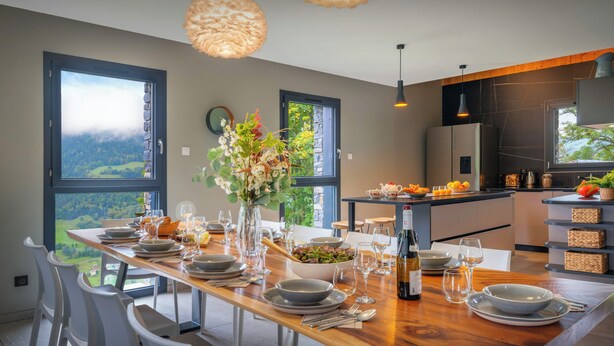 A modern dining area with a wooden table set for a meal, large windows offering a view of the outdoors, and a kitchen in the background.