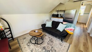 Ett kompakt vardagsrum med en svart soffa, ett litet träbord och en mönstrad matta. En mikrovågsugn och ett kylskåp syns i bakgrunden.