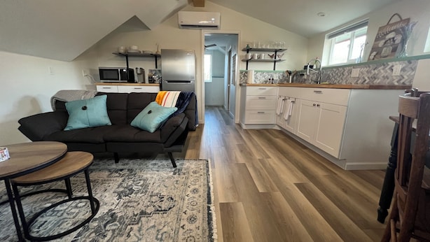 Ett kompakt bostadsutrymme med ett litet kök, en soffa med kuddar, ett matbord och en mönstrad matta.