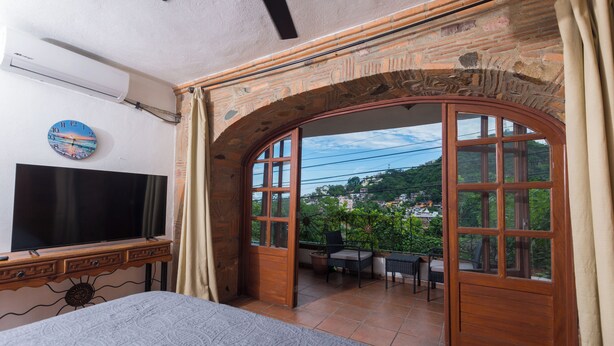 Une chambre dotée d’une télévision, d’un lit, d’une fenêtre donnant sur un espace verdoyant et d’un balcon avec une table et des chaises.