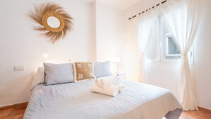Un dormitorio con cama, almohadas, un espejo montado en la pared y una ventana con cortinas.