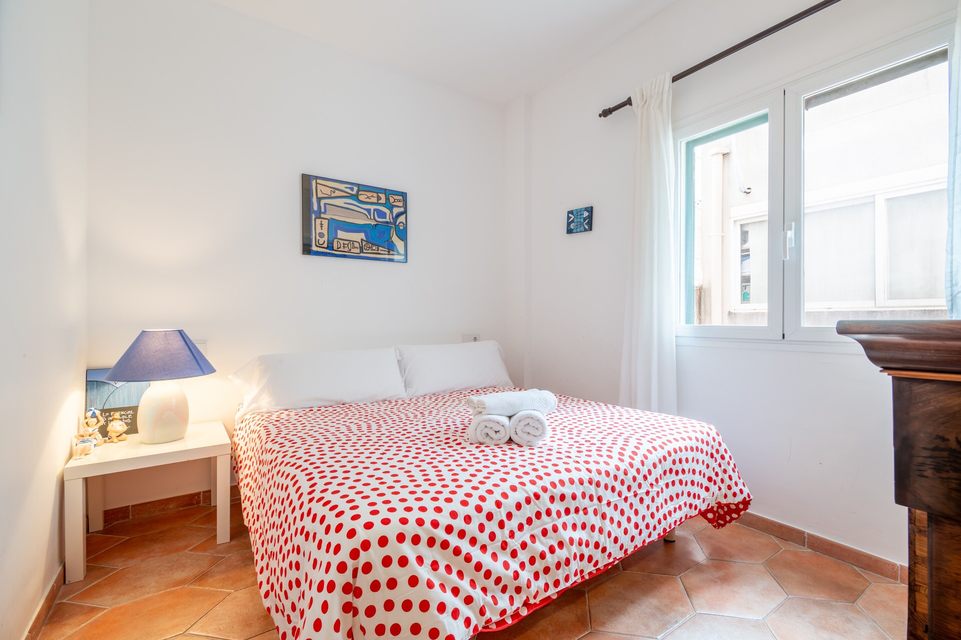 Un dormitorio con cama, mesita de noche, lámpara, ventana y un cuadro en la pared.