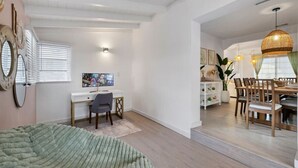 A room with a desk, chair, and computer, a dining area with a table and chairs, and a bed with a green blanket.
