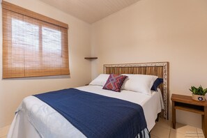 Une chambre à coucher avec un lit, une fenêtre avec des stores, une table de chevet avec une plante et une tête de lit ornée d’un motif décoratif.