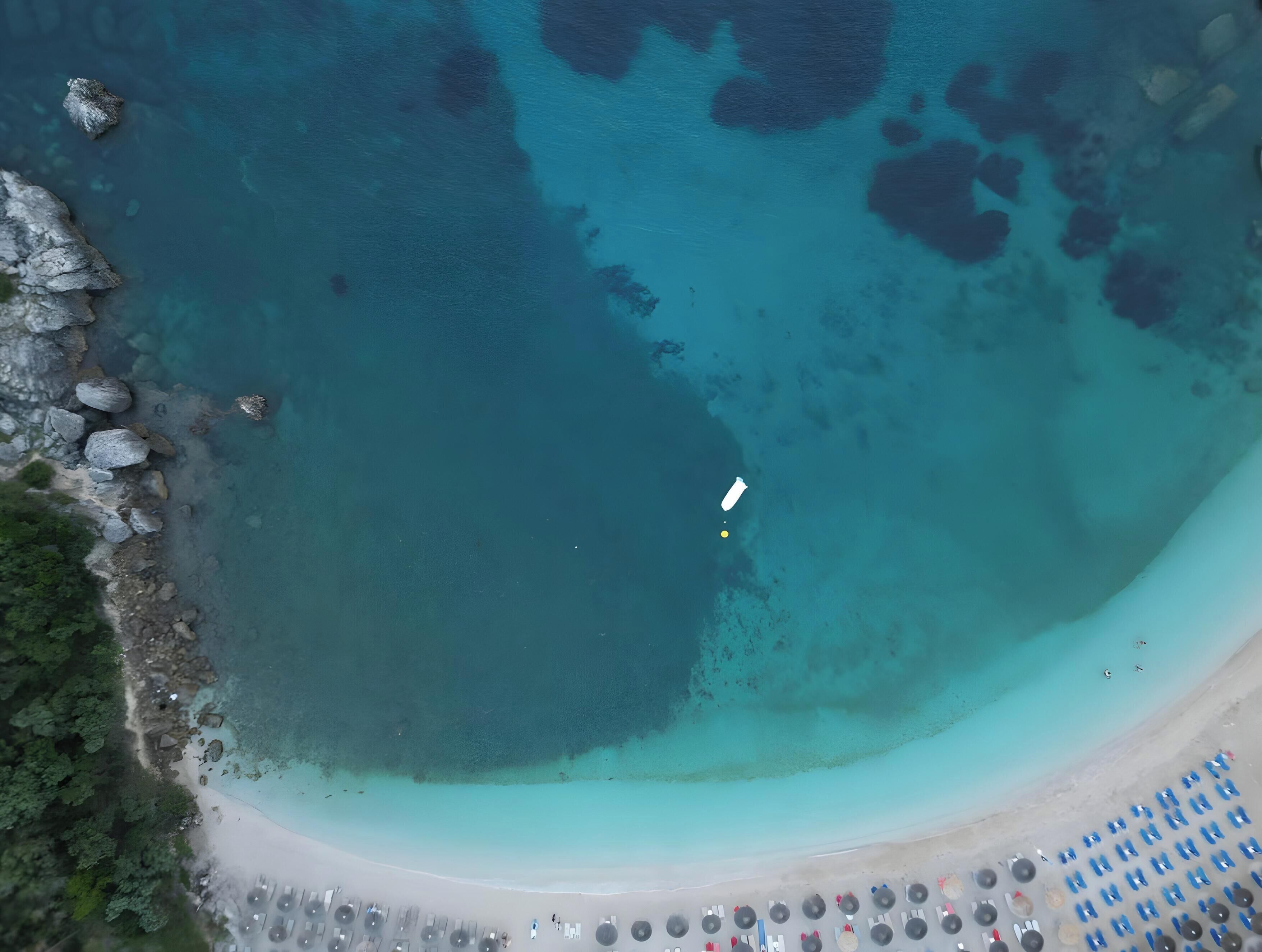 Luftaufnahme eines Strandes mit klarem türkisblauem Wasser, einem weißen Boot und einer Reihe von Strandsonnenschirmen.