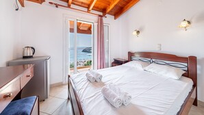 Une chambre avec un plafond en bois, un lit avec du linge de maison blanc, un bureau avec une bouilloire et une vue sur la mer par une porte vitrée.