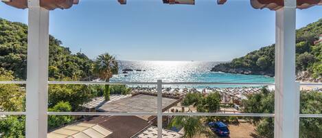 A view from a covered balcony overlooking a beach with a clear sky, a parking area, and lush greenery.