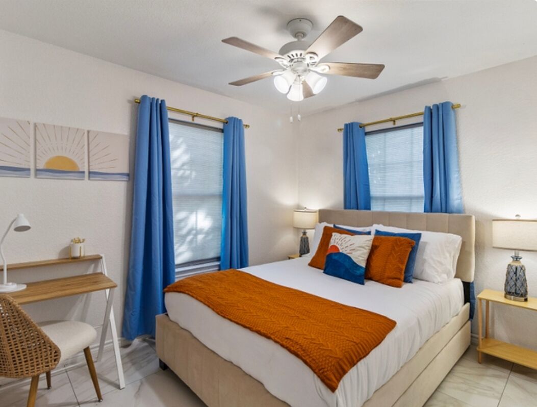 Ein Schlafzimmer mit Bett, Schreibtisch, Stuhl, Deckenventilator und Vorhängen.